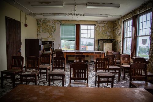 Abandoned classroom with wooden chairs, peeling paint, and large windows, creating a desolate atmosphere.