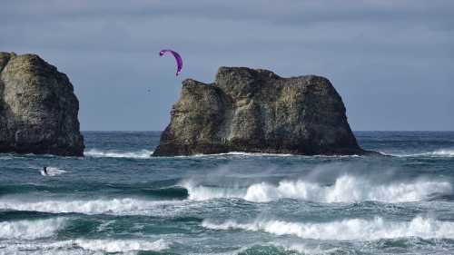 A kitesurfer glides over waves near rocky cliffs under a cloudy sky.