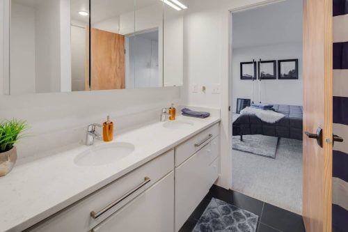 Modern bathroom with double sinks, mirrors, and a doorway leading to a bedroom. Neutral colors and sleek design.