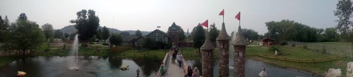 A panoramic view of a park featuring a pond, fountain, castle-like structures, and green trees in the background.