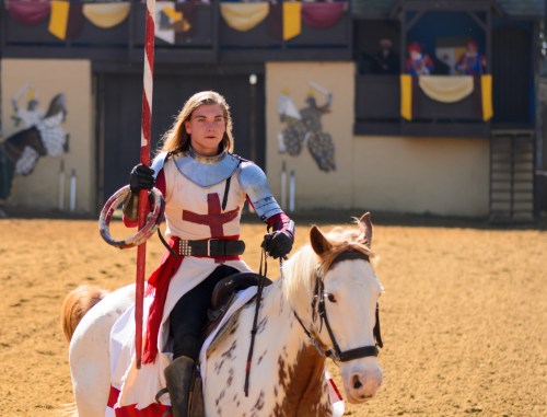 A knight in armor with a red cross rides a white horse, holding a lance, in a medieval tournament setting.