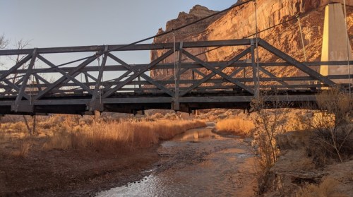 A wooden bridge spans a shallow creek, surrounded by rocky cliffs and dry vegetation in warm evening light.