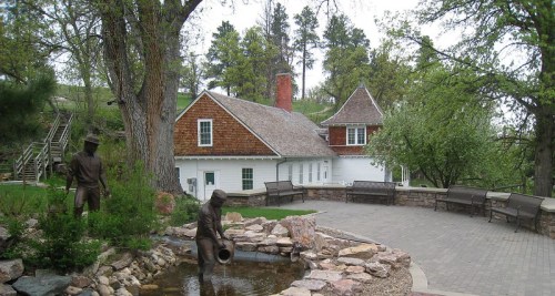 A charming house with a shingled roof, surrounded by trees and a small pond with a statue of a person by the water.