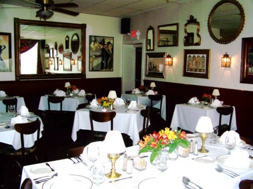 Cozy restaurant interior with white tablecloths, elegant decor, and floral centerpieces, featuring mirrors and artwork on the walls.