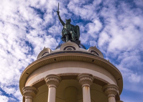 A statue of a warrior holding a sword atop a circular building, with a blue sky and clouds in the background.