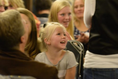 A smiling young girl in a striped shirt sits among a crowd, looking up with joy at someone speaking.