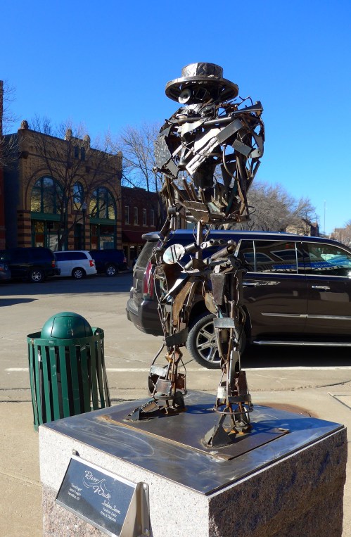 A metal sculpture of a humanoid figure stands on a pedestal in an outdoor setting, with buildings in the background.