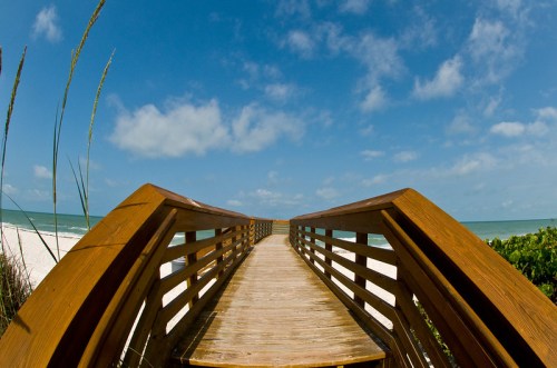 A wooden boardwalk leads to a sandy beach under a blue sky with scattered clouds.