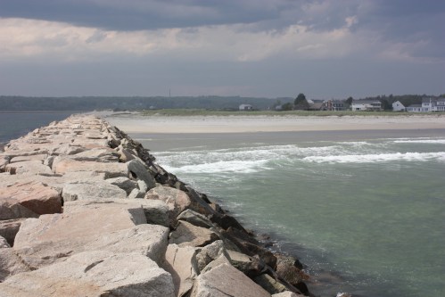 A rocky jetty extends into the water, with a cloudy sky and a distant shoreline visible in the background.