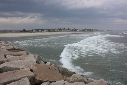 A rocky shoreline with waves crashing, leading to a beach and a distant coastal town under a cloudy sky.