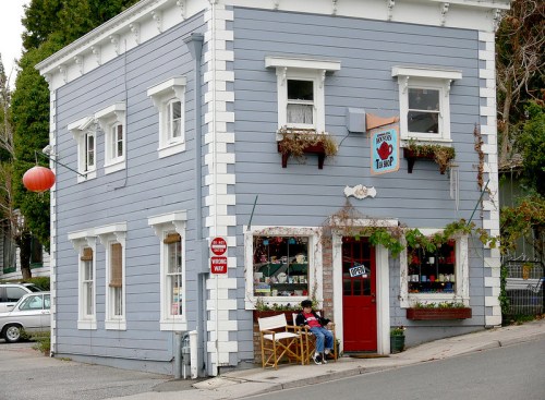 A charming blue building with a red door, featuring flower boxes and an "Open" sign, sits on a sloped street.