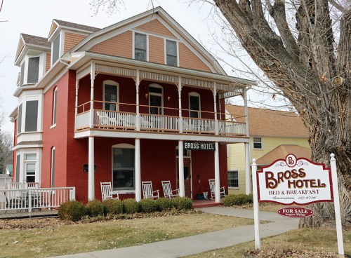 A red brick building with a porch, labeled "Bross Hotel," surrounded by grass and trees, with a "For Sale" sign.