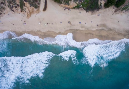 Aerial view of a sandy beach with waves crashing, surrounded by cliffs and scattered beachgoers.