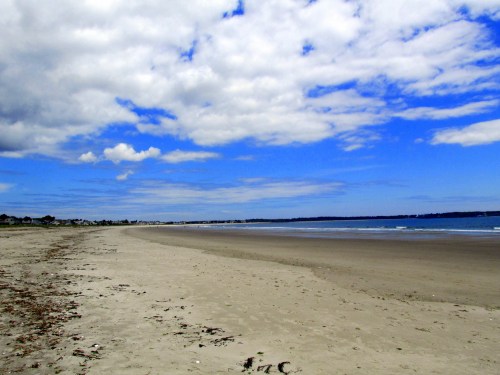 A serene beach scene with soft sand, gentle waves, and a blue sky dotted with clouds.