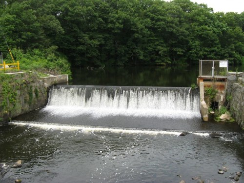 A small dam with water cascading over it, surrounded by lush green trees and a calm river.