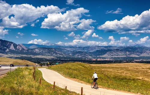 A cyclist rides along a paved path with mountains and a blue sky filled with clouds in the background.