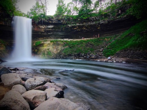A serene waterfall cascades into a rocky pool, surrounded by lush greenery and a cliff, with people in the background.