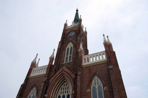 A tall brick church tower with a clock and spire, set against a cloudy sky.