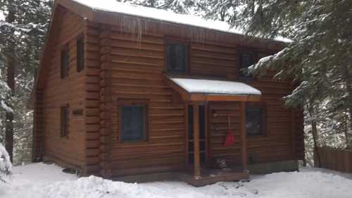 A cozy log cabin surrounded by snow-covered trees, with a small porch and icicles hanging from the roof.