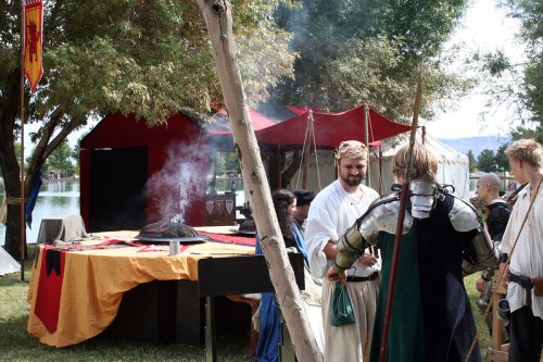 A medieval fair scene by a lake, featuring costumed participants, tents, and a cooking area with smoke rising.