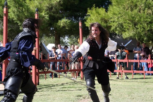 Two men in historical costumes duel with swords in a grassy area, while an audience watches in the background.