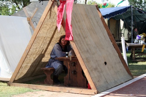 A person sits at a wooden table inside a tent made of fabric and wood, surrounded by other tents in a grassy area.