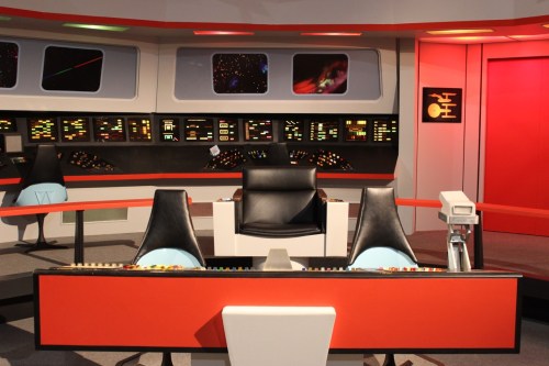 A futuristic control room with black and red decor, featuring a central chair and various colorful control panels.