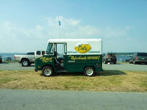 A vintage ice cream truck parked by a lake, advertising soft frozen lemonade with a bright yellow logo.
