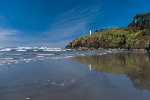 A scenic beach with gentle waves, a lighthouse on a rocky cliff, and a clear blue sky reflecting on the wet sand.