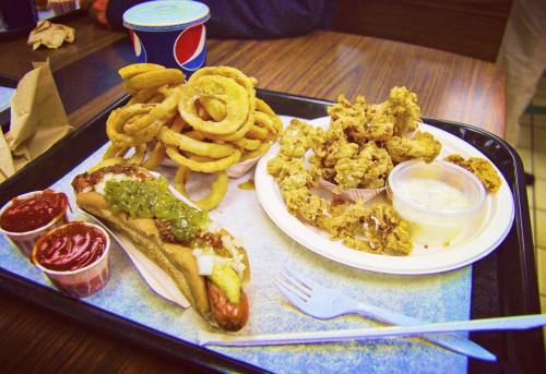 A tray with a hot dog topped with condiments, onion rings, fried chicken pieces, and dipping sauces.