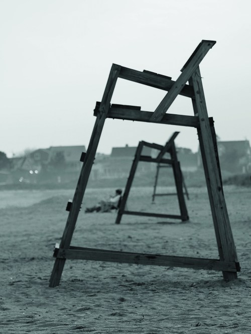 Three wooden structures stand on a sandy beach, with a hazy background of houses and a person lying down.