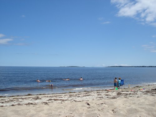 A sunny beach scene with people swimming in the water and two individuals standing on the shore.