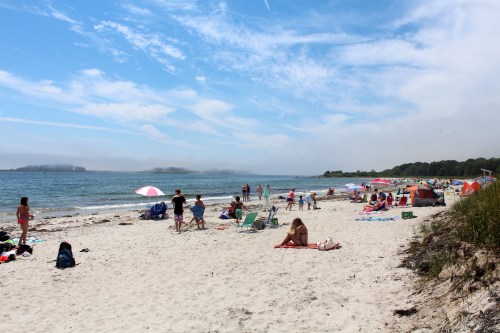 A sunny beach scene with people relaxing on the sand, colorful umbrellas, and calm water in the background.