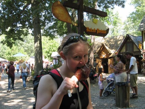 A woman bites into a large turkey leg at a festival, surrounded by trees and people enjoying the event.
