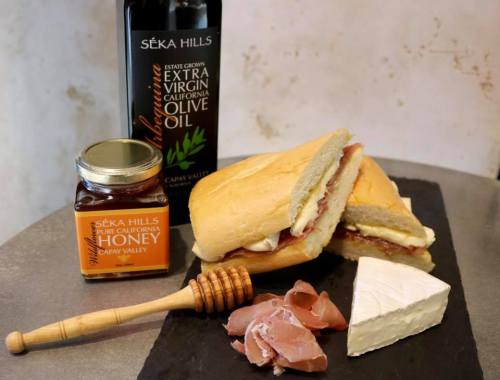 A rustic display featuring a sandwich, honey jar, olive oil bottle, cheese, and prosciutto on a slate board.