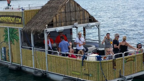 A party boat with a thatched roof, people enjoying themselves, and tropical decorations on a sunny waterway.
