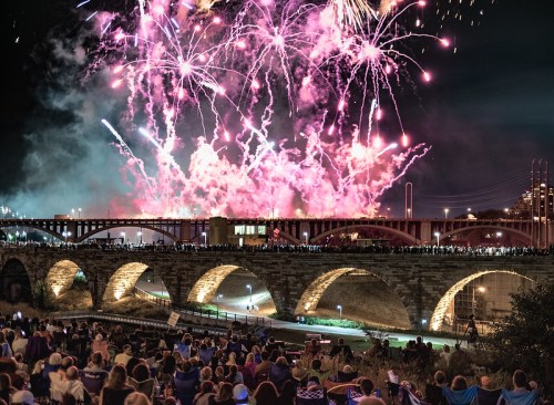 Crowd watching vibrant fireworks display over a bridge at night, with colorful bursts lighting up the sky.