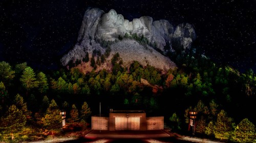 Mount Rushmore illuminated at night, surrounded by trees under a starry sky.