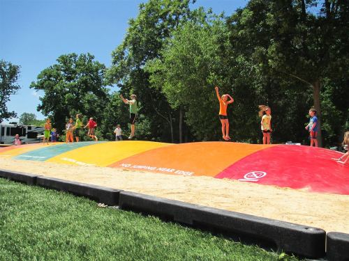 Children in colorful shirts jump on a large inflatable bounce pad in a park surrounded by trees.