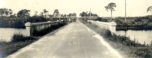 A vintage photo of a bridge over a waterway, surrounded by trees and open fields, with a dirt road leading into the distance.