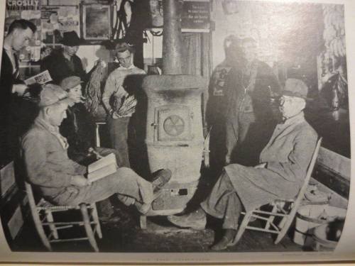 A group of people gathered around a stove in a cozy, vintage setting, engaged in conversation and reading.