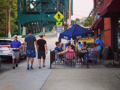 People walking along a sidewalk near a restaurant with outdoor seating, under a bridge on a sunny day.