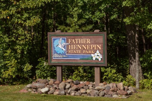 Sign for Father Hennepin State Park, featuring a blue bird and surrounded by rocks, set against a green forest backdrop.