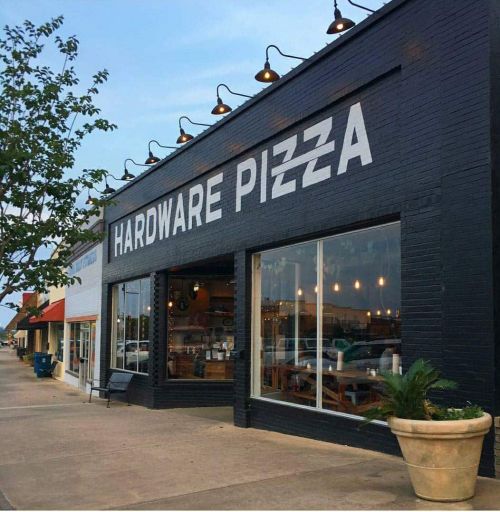 Exterior of Hardware Pizza, featuring a large sign, windows, and outdoor seating in a commercial area.