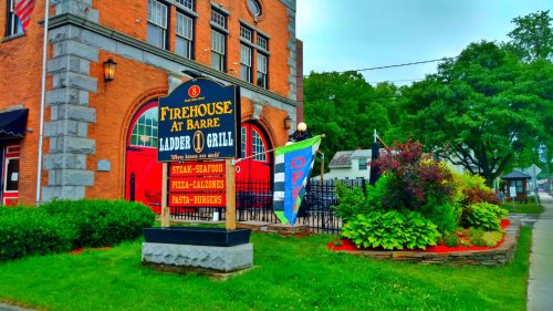 Sign for "Firehouse at Barre Ladder Grill" featuring food options like steak, seafood, and burgers, with colorful landscaping.
