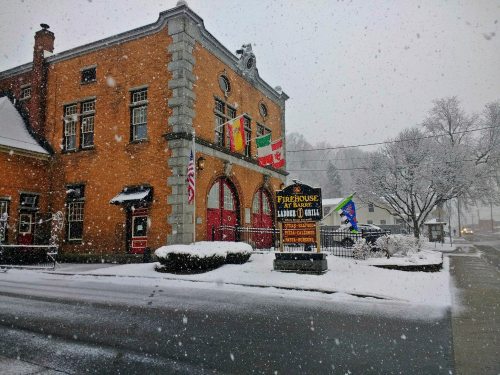 Historic firehouse in snowy weather, featuring flags and a sign, surrounded by a winter landscape.