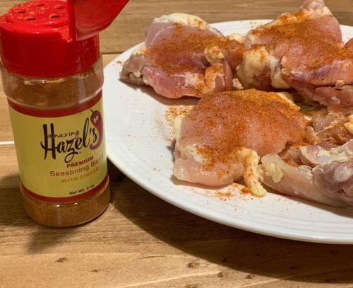 A plate of raw chicken thighs seasoned with a spice blend next to a jar of Amazing Hazel's seasoning.