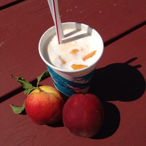 A cup of creamy drink with ice and fruit, accompanied by two fresh apples on a wooden surface.