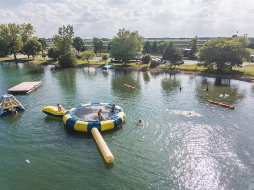 Aerial view of a lake with inflatable water toys and people swimming, surrounded by trees and a clear sky.