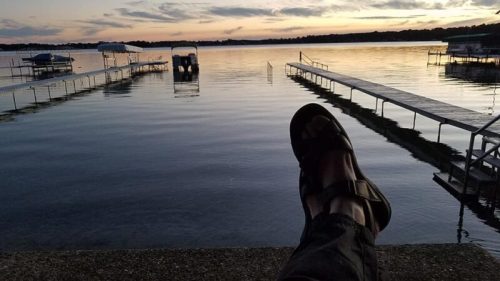A person’s foot in sandals rests on a dock, overlooking a calm lake at sunset with boats and piers in the background.